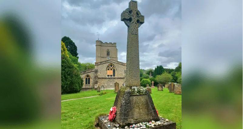 War memorial, with pebbles and a poppy wreath at its base, surrounded by lawn, with church and gravestones in background.