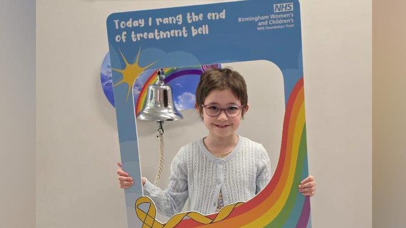 Dulcie has short brown hair and glasses. She is holding a rainbow sign that says "today I rand the end of treatment bell". There is a silver bell on the wall behind her