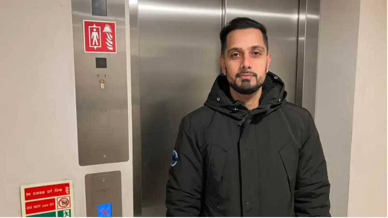 Shazan Qureshi wearing a black coat and standing in front of the broken lift