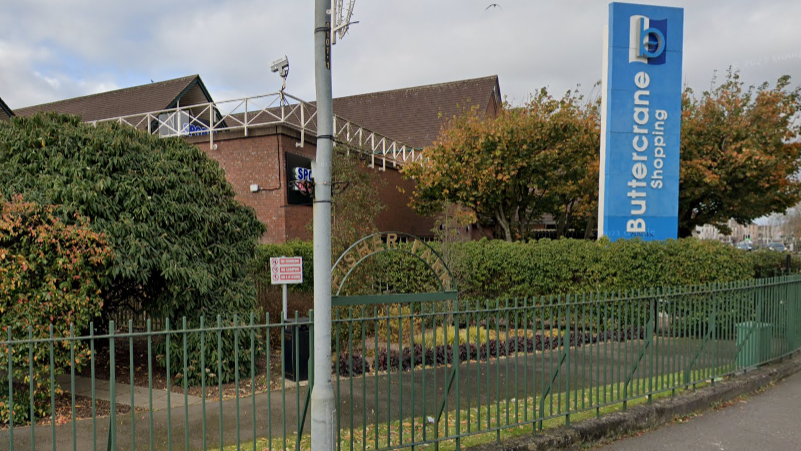 A wide view of Fisher Park, a small section of parkland which skirts around the edge of Buttercrane Shopping Centre. The park is fenced off with green iron railings. There is a small path lined with shrubs. A large blue sign for the shopping centre is in the background.
