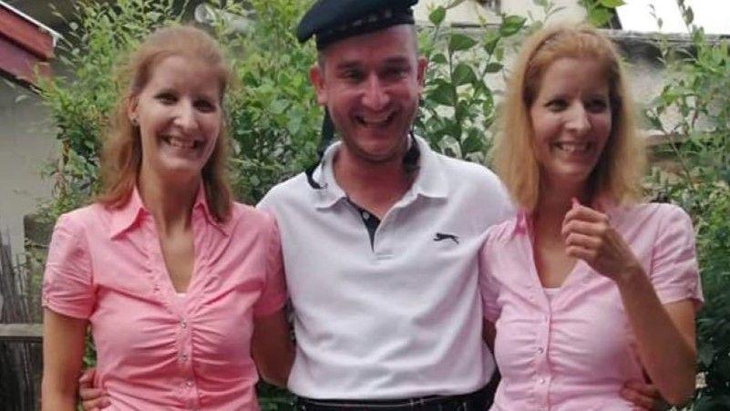 Two twin sisters wearing pink tops, either side of a man in a white top.