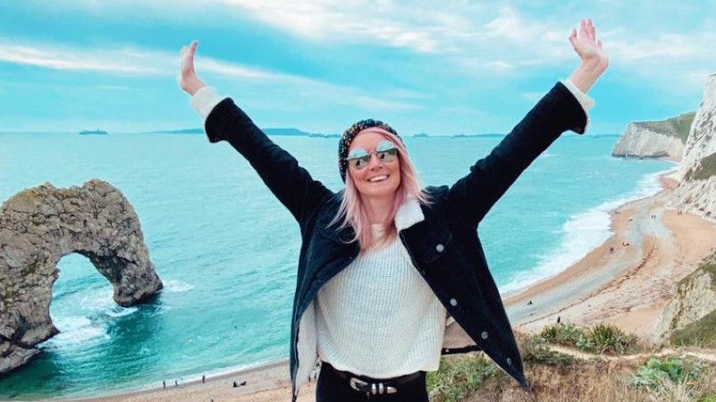 Clare Elgar, who has MS, stands on a beach, smiling at the camera, arms raised in the air