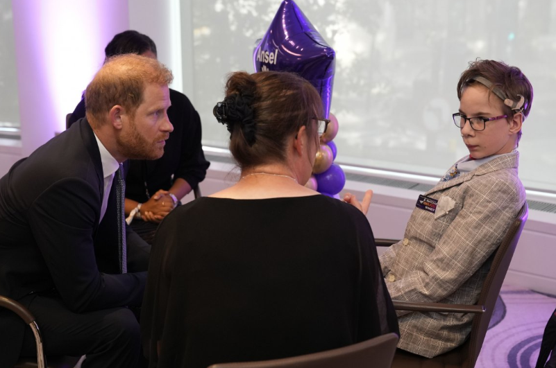 Prince Harry, with balding red hair and a trimmed beard, sits in a black suit opposite a boy in a grey checked suit with a hearing device on his head. A woman in a black dress sits across from them with her back to the camera. There is a purple star-shaped balloon in the background with the name Ansel on it.