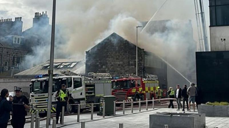 Two fire engines beside a burning brick building. Smoke comes from the roof as water is sprayed from a hose and people watch on.