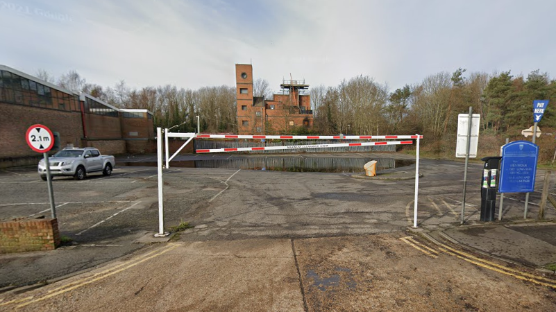 A Google streetview image of the entrance of Henwood car park. The car park is empty bar from one grey car and there is a red and white barrier at the entrance.. 