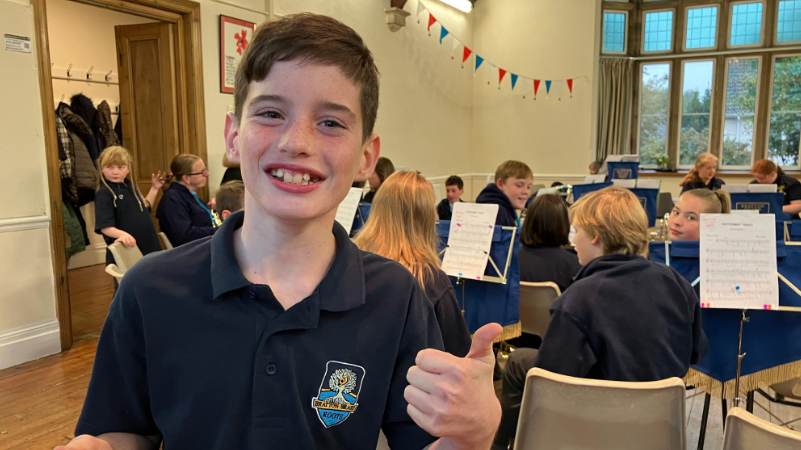 Joshua is wearing a navy blue shirt and giving thumbs-up. He is smiling. He is standing in a practice room with other brass players in the background.