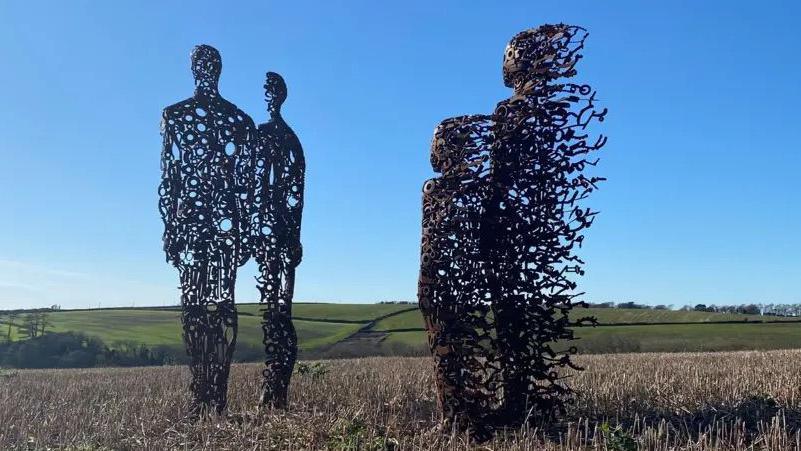 Four statues facing away from the camera, made of scrap metal, in a field. In the background are green fields and a bright blue sky.