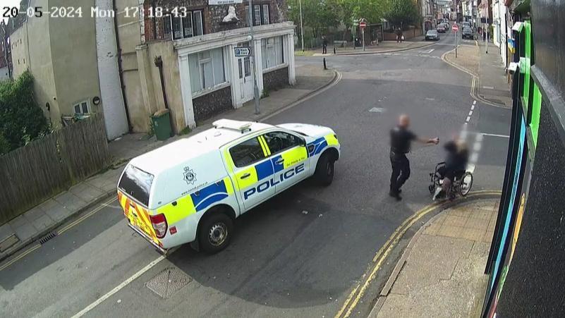 A police truck parked in the middle of King Street, Great Yarmouth, with the police officer, arm extended, deploying PAVA spray on Michael McQueen, who is in his wheelchair on the white line of the junction of St Peter's Road. Behind the police van is the Old White Lion pub, which is converted into flats, and to the right of the image is the frontage of the Londis convenience store. In the background is King Street looking towards Great Yarmouth town centre.