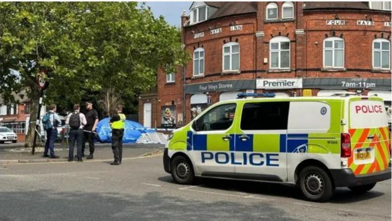 A police van to the right of the frame parked near a cordon on Reedswood Lane in Walsall while four men speak to each other under a tree.  