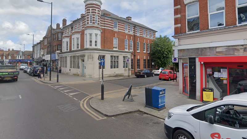 The junction of two roads. There are cars parked along both sides of both roads.