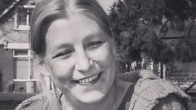 A black and white picture of Dawn Sturgess smiling at the camera, wearing a t-shirt, cardigan and a necklace. She is sitting on the grass in front of a house and a tree.