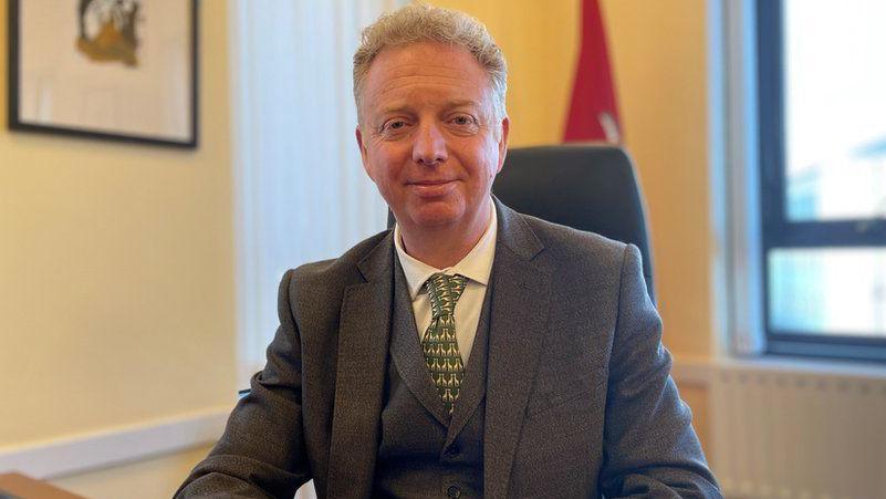 Alex Allinson sitting at his office desk, with an Isle of Man flag standing behind him. He has short, wavy blond hair and is wearing a grey suit with a waistcoat and a pattered green tie.