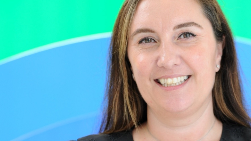 A headshot of Hannah Coffey smiling at the camera. She has long, brown hair. The picture has been taken against a bright blue and green background.