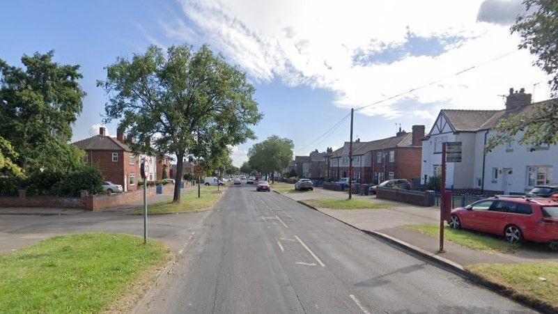 A google maps streetview image of Fryston Road. It is a fairly average residential street. Cars are driving up and down the road. The houses on either side of the street are quite large. There are cars parked near the houses, on driveways, and on the public grass verges outside. 