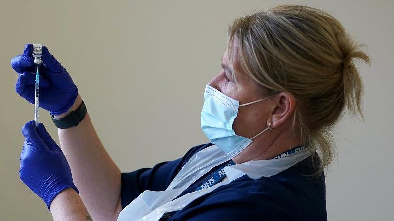 A nurse preparing a vaccine. She is wearing a blue uniform and NHS lanyard and she has blue plastic gloves, a white plastic apron and a mask. Her hair is tied back.