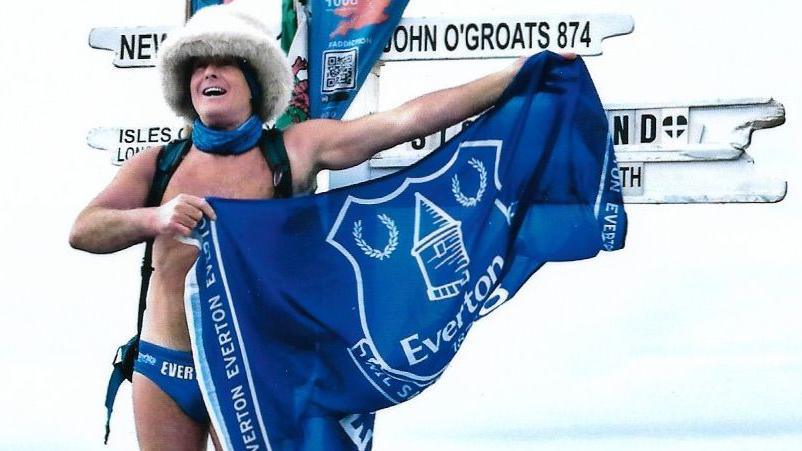 Speedo Mick, wearing a white furry hat, a rucksack and blue swimming trunks, holds out a blue Everton flag in front of the signpost at Land's End