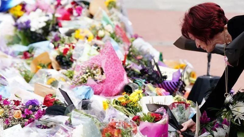 Sharon Osbourne is on the right of the shot bending down to lay flowers in memory of her husband. She has dark red short hair and is dressed in black. In front of her, there is a sea of floral bouquets laid by fans.
