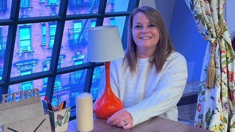 A lady sitting at a table in front of a window. She is smiling and looking at the camera, wearing a cream jumper and she ahs mousy brown hair to her shoulders. There is an orange lamp in front of her on the table. 