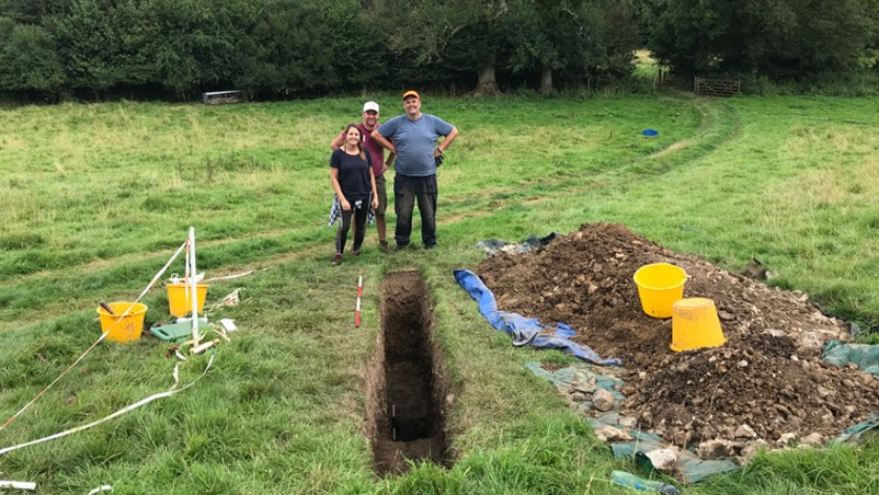 The Bournemouth University excavation team (L-R) Sarah Elliott, Harry Manley and Mark Johnson. Sarah is wearing black leggings, a black top and a checked shirt round her waist, Harry is wearing a red t-shirt and green shorts whilst Mark is wearing a blue t-shirt and black trousers.