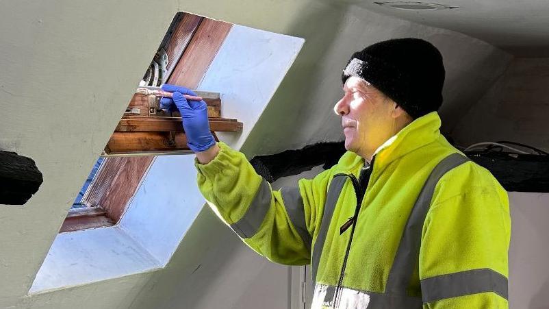 A man is stood inside a room while painting a window fame. He wears a woolly hat, blue latex gloves and a thick yellow high visibility jacket. 