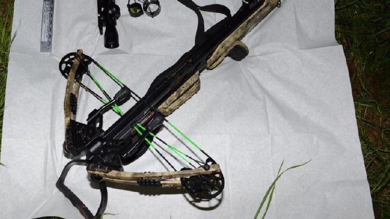 A crossbow, which appears to have a camouflage-like trim and black metallic holder, lying on a white sheet on top of grass