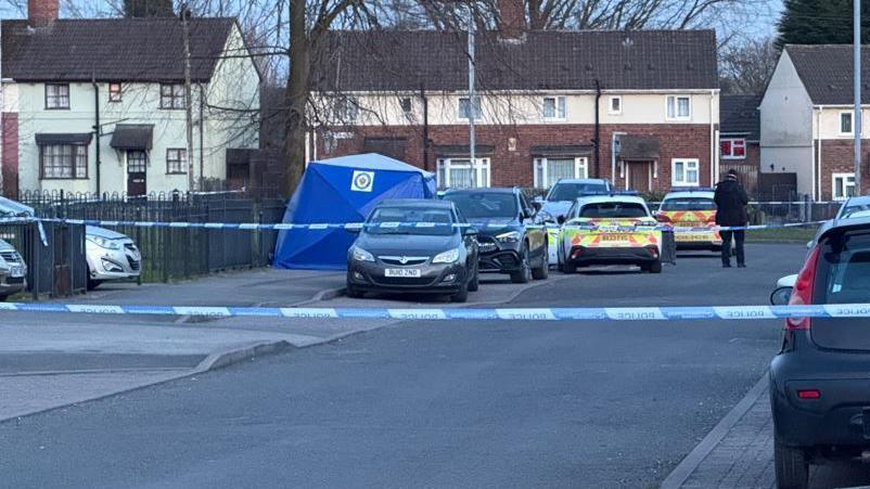 A residential street that is cordoned off with police tape. There are two police cars in the background and an officer standing, with their back to the camera, near the tape