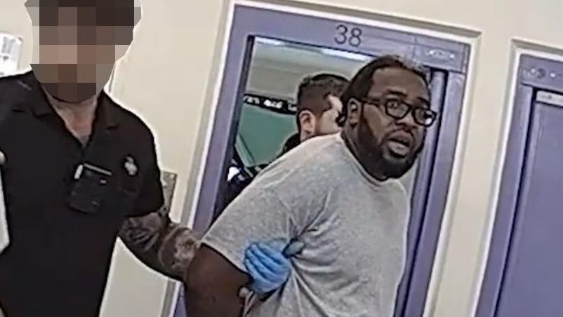 An image of a man wearing glasses with his arms behind his back, being detained by a prison officer