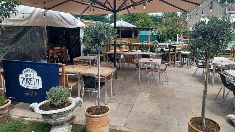 An outside image of the pub garden with some umbrellas up and lights hanging. There are plants around the pathed area.