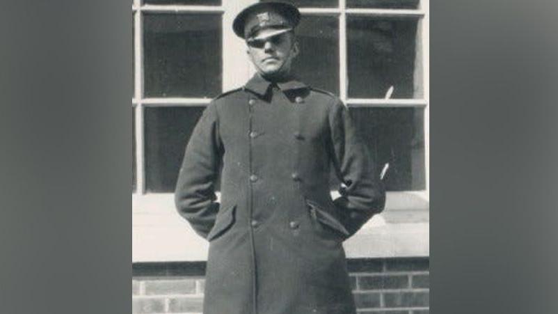 A man standing with his hands behind his back. He is wearing a long black coat and a police hat. He is standing in front of a building.