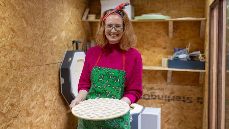 Sarah Cook is standing in a shed holding a tray with ceramic hearts on it towards the camera. She is smiling. She is wearing a pink jumper and green apron.
