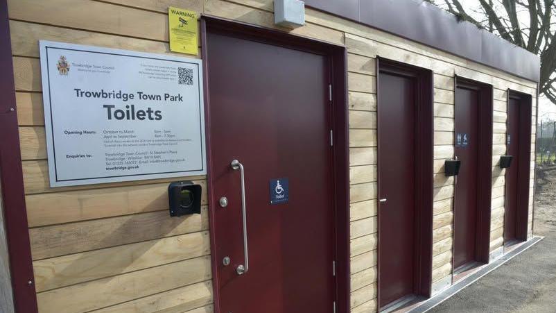 Four red doors in a portable-style wooden building with a sign which says "Trowbridge Town Park Toilets"