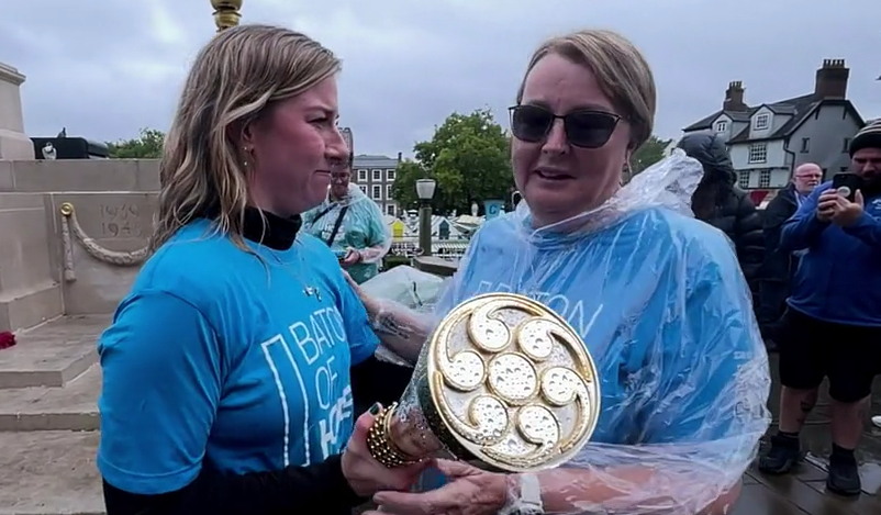 Two women standing side on to the camera, wearing blue tshirts, both hold a gold and silver baton - the top end of which is facing the camera. The woman on the right is wearing sunglasses and her tshirt is covered by a clear plastic raincover. The woman on the left is looking at the other woman. Behind them are people taking photos on mobile phones, the Norwich war memorial and Norwich market stalls