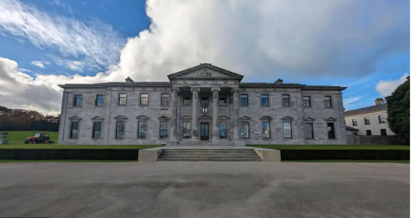 A large, historic looking house - the Ballyfin Demesne - is seen from the front on. It has stone steps leading up to a front door surrounded by columns. There is a cloudy sky in the background. 