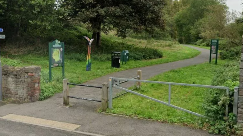 The entrance to a park with a low gate, two information boards and a path leading away between trees.