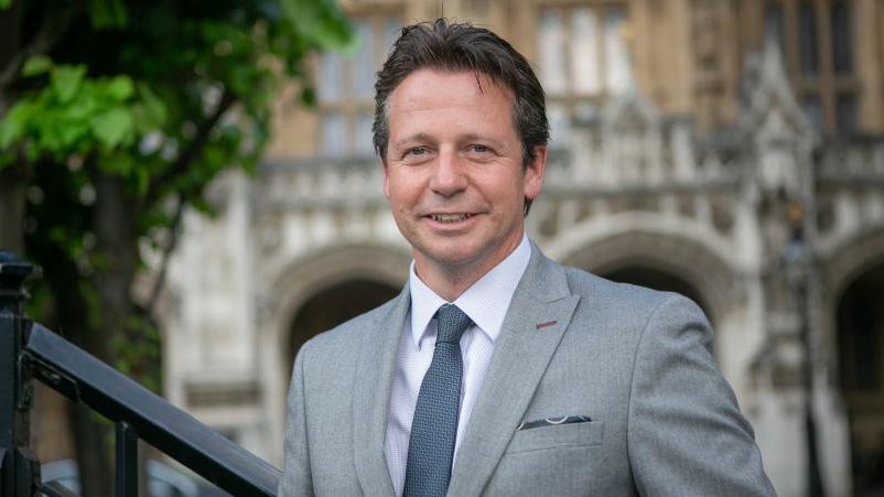 A man with short dark hair, wearing a grey suit, pale shirt and blue tie smiles at the camera.