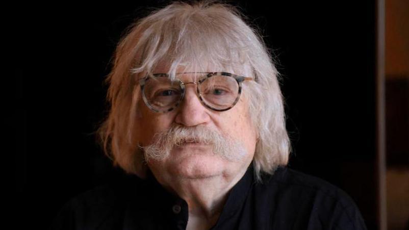 Sir Karl Jenkins has a black shirt on and has tortoise-shell round-rimmed glasses. He has a white moustache and white shoulder length hair