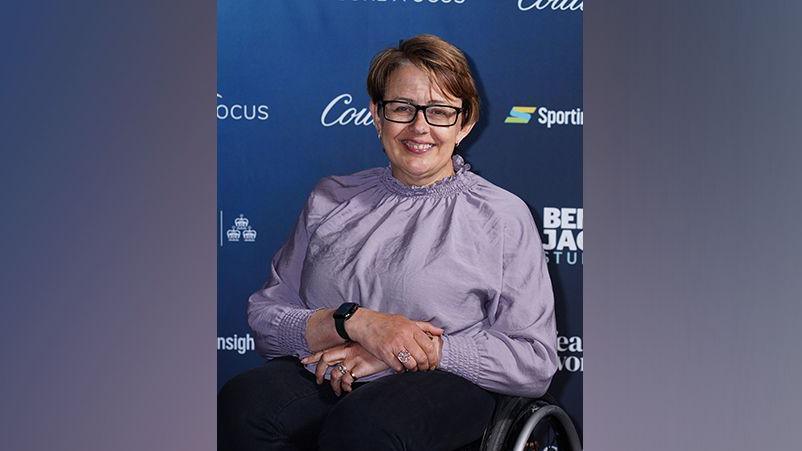 Baroness Tanni Grey-Thompson looking direct to camera. She is wearing a lilac top, with frilled collar and cuffs, black trousers, and dark framed, rectangular glasses. She is seated in a wheelchair. The top of one wheel is visible on the right of the image. Her hands are folded on her lap. She is posing in front of an advertising hoarding, which has the names of several brands printed on it.