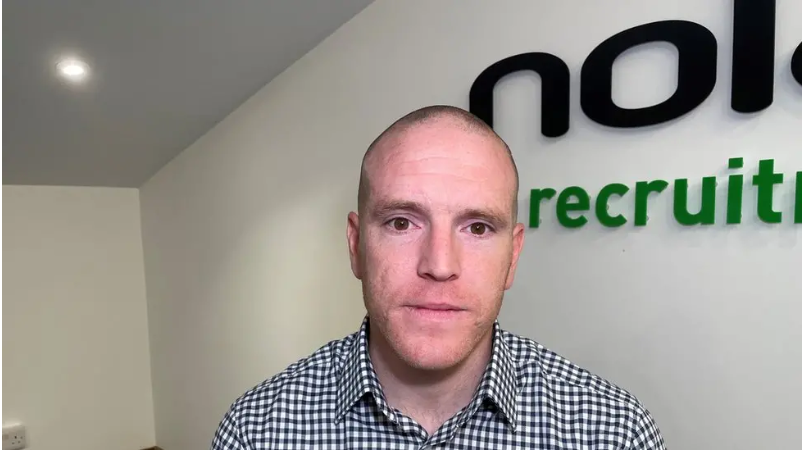 A man in a chequered white and black shirt is standing in front of a white wall with the black and green company logo in the background