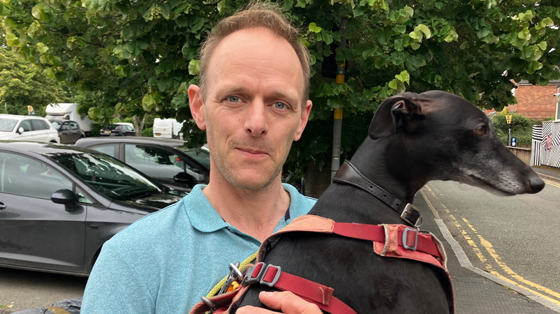 A man holding a black dog in his arms looks at the camera