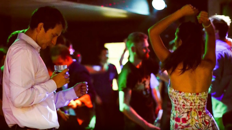 A man and a woman dancing in a nightclub setting, with multicoloured disco lights. The man has short brown hair is wearing a pink shirt and holds a glass, and the woman has long brown hair is wearing a strapless white dress, patterned with pink and green flowers.