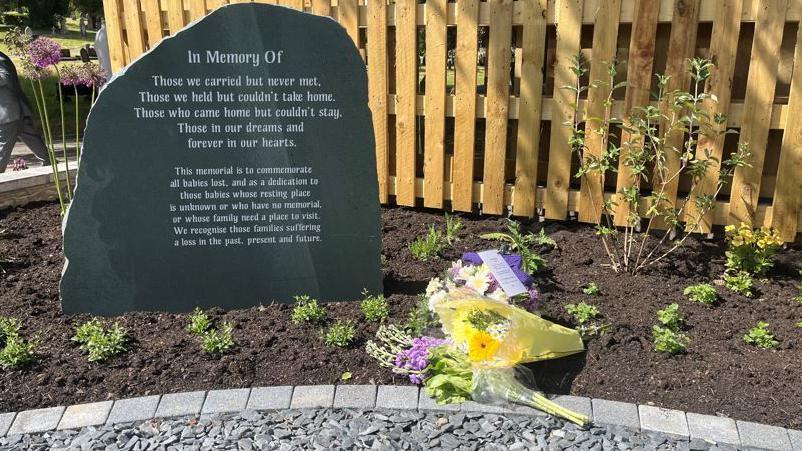 The black memorial stone is on soil surrounded by three bouquets of flowers and small plants. There is a wooden fence behind. The memorial reads: In memory of those we carried but never met, those we held but couldn't take home, those came home but couldn't stay, those in our dreams and always in our hearts.