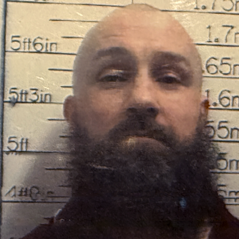 Michael Ross who has a bald head and a big brown, bushy beard. He is standing in front of a height chart and is looking at the camera in what appears to be a police photograph.