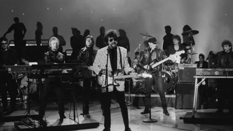 A black and white image of Jeff Lynne fronting the original line-up of the Electric Light Orchestra, playing live in 1972. He has his trademark sunglasses on and is wearing a light coloured suit jacket and black trousers, and is holding an electric guitar.