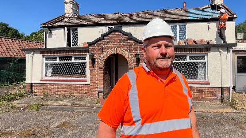 Kiel Barber standing outside the Decoy Tavern in Fritton. He is wearing a white hard hat and an orange high visibility tee-shirt, with silver stripes on the front. He is standing in front of the pub, which has some of the roof tiles removed as the building is being demolished. It is a cream coloured building with a symmetry of bay windows and first floor windows, and an extension to the right of the image.