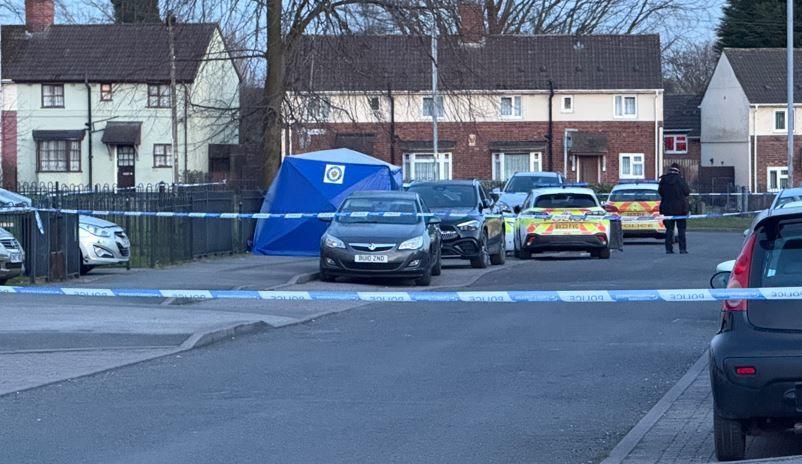 A residential street that is cordoned off with police tape. There are two police cars in the background and an officer standing, with their back to the camera, near the tape