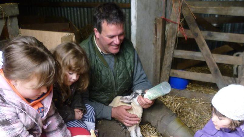 A man and three young girls seated in what appears to be a barn, with straw on the ground and a five-barred gate visible to the right. He is wearing a dark green gilet and bottle feeding a lamb. To his left are two young girls, his nieces, one is wearing a pink and grey checked fleece, the other a dark jacket. On his right can be seen the head and shoulders of another niece, who is wearing a purple jacket and white hat.