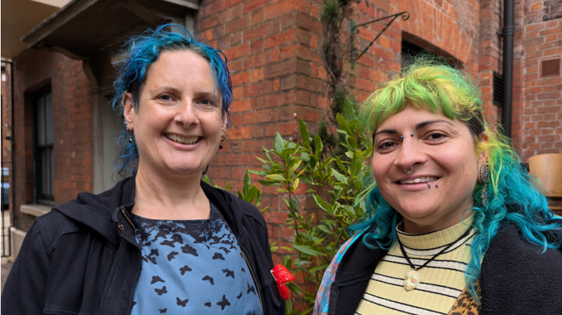 Two women looking at the camera - one on the left wears a navy jacket and has blue colour in her hair - the one on the right wears a stripey top and has vivid blue and green colour in her hair