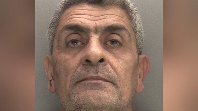 A mugshot of a man, staring at the camera, in front of a grey background