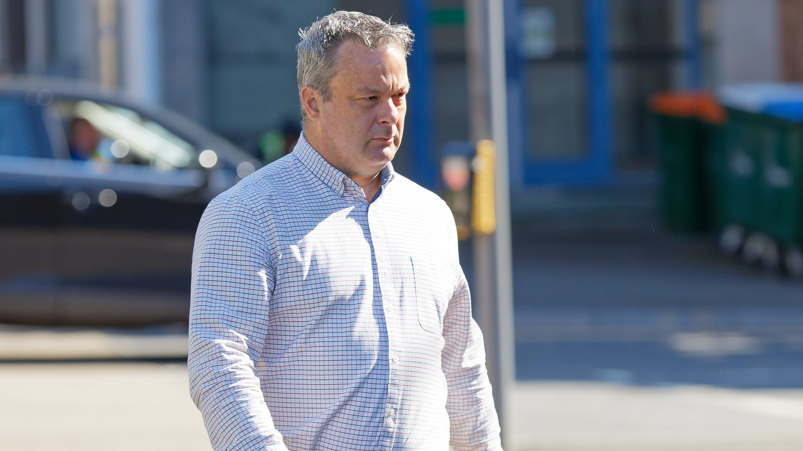 A man with grey hair walking. He has a white checked shirt on.