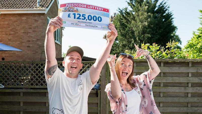 Jamie Holden wearing a white and green cap, wearing a white T-shirt and sporting a tattoo on his right upper arm. He is holding a large cheque above his head. Laura Fullthorpe has long dark hair and is wearing a pink top and white T-shirt. She is smiling broadly and pointing upwards. There is a wooden fence behind them with a hedge behind that.